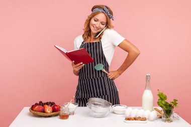 Şef aşçı ya da apronlu fırıncı, fırın malzemeleriyle dolu masada kek pişiriyor, cep telefonuyla konuşuyor, müşteri siparişi alıyor, defterine bakıyor. Pembe arka planda kapalı stüdyo çekimleri.