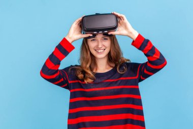 Dalgalı saçlı, çizgili süveter giyen, sanal gerçeklik kulaklığı deneyimi olan, 3 boyutlu simülasyon, yüksek teknoloji, mutlu yüz ifadesi olan bir kadın. Mavi arka planda kapalı stüdyo çekimi.