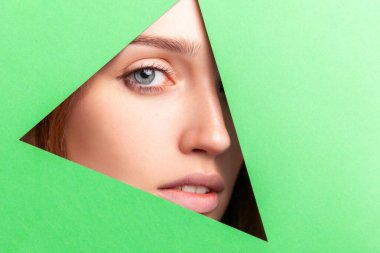 Makyajsız genç, çekici bir kadın yüzü, kameraya bakan bir kadın, üçgen, yeşil kağıt deliğinden bakan bir kadın. Cilt bakımı ve güzellik anlayışı. Stüdyo içi çekim.