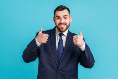 Sakallı, resmi stil takım elbiseli yakışıklı, dişlek gülüşlü kameraya bakan, baş parmağını kaldıran, neşeli bir adam, harika haberler duyuyor. Mavi arka planda kapalı stüdyo çekimi.