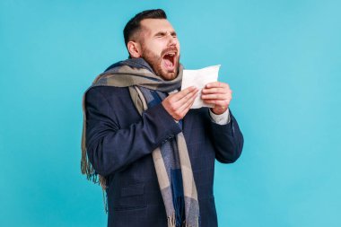 Öksürme ya da hapşırma. Eşarba sarılı hasta bir adam hapşıran doku, öksüren, kendini kötü hisseden, hasta ya da grip olan, alerji ya da mevsimsel grip belirtileri gösteren. Mavi arka planda kapalı stüdyo çekimi