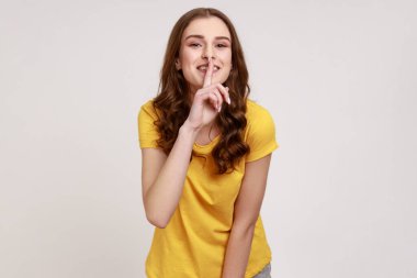 Lütfen, sessiz olun. Olumlu genç kız sessiz hareketlerle sessiz duruyor, parmaklarını dudaklarına dayayıp kameraya gülümsüyor, sır tutmak istiyor. Gri arka planda izole edilmiş kapalı stüdyo görüntüleri.