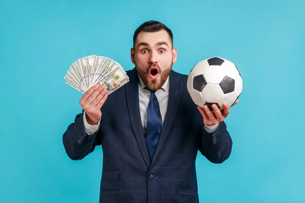 サッカーボールを保持する公式スタイルのスーツを身に着けているひげを剃った男と100ドルの紙幣 賭けと勝利 口を開いておく 青い背景に隔離された屋内スタジオショット — ストック写真