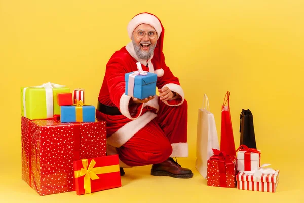 サンタのクラスの衣装を着た白髪の老人は 現在の箱を包んだ近くに座って 贈り物をし 新年を祝いました 黄色の背景に隔離された屋内スタジオショット — ストック写真