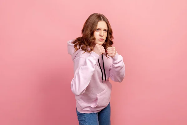 フーディで怒っている10代の女性の肖像画は パンチする準備ができて 拳や激怒した表情で立っている ピンクの背景に隔離された屋内スタジオショット — ストック写真