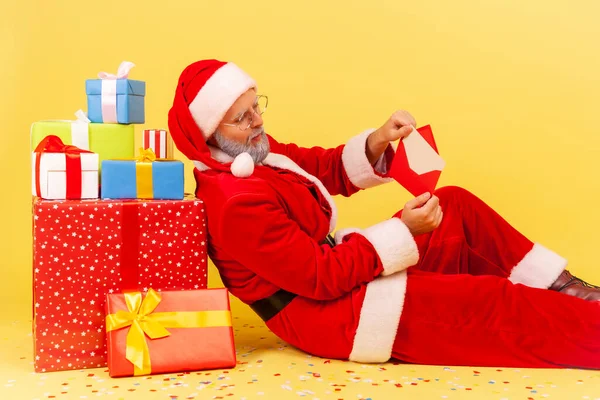 Side view of elderly man with gray beard in santa claus costume sitting on floor near present boxes, reading letter with Christmas congratulations. Indoor studio shot isolated on yellow background.