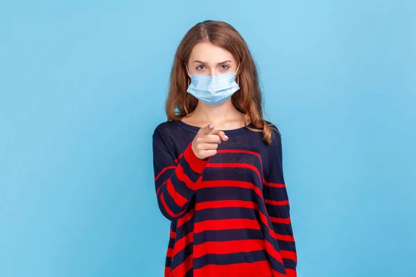 Hey you. take care of you health. Portrait of serious woman wearing sweater and surgical medical mask standing, pointing and looking at camera and blame. Indoor studio shot isolated on blue background