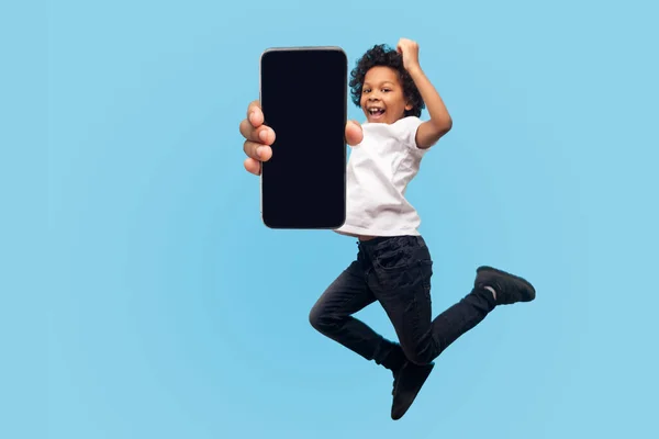 完全な長さ 活気のある活気のある小さな男の子の幸福とジャンプし 携帯電話の画面を表示し 驚きの勝者の顔を 子供を飛んで インスピレーションを感じている 屋内スタジオ撮影青の背景 — ストック写真