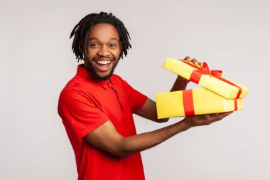 Dişli tebessümlü mutlu bir adam kırmızı günlük tişört giyen, el ele tutuşmamış hediyeler taşıyan, hediyeyle tatmin olan rastalı saçları var. Gri arka planda izole edilmiş kapalı stüdyo görüntüleri.