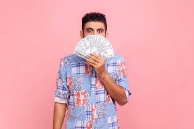 Sakallı, mavi gömlekli çekici bir adamın portresi çok para gösteriyor, ağzını kapatıyor, büyük gözlerle kameraya bakıyor. Pembe arka planda kapalı stüdyo çekimleri.