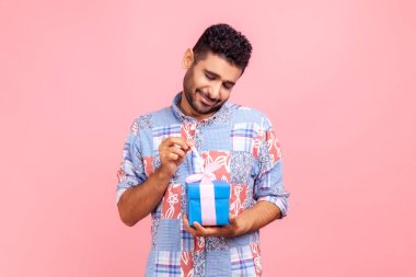 Mutlu sakallı adam hediye kutusunu açıyor, bayramı kutlamak için doğum günü hediyesini açıyor, kurdele çekiyor, hediyeye bakıyor, mavi bir gömlek giyiyor. Pembe arka planda kapalı stüdyo çekimleri.