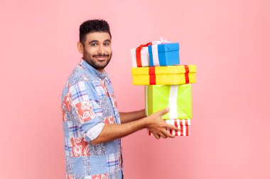 Sakallı bir adamın elinde hediyelerle kameraya bakarken, hoş bir gülümsemeyle, bayramı kutlarken, mavi bir tişört giyerken. Pembe arka planda kapalı stüdyo çekimleri.