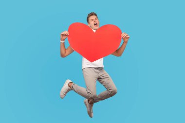 Aşkta mutlu bir adam zıplar ve büyük kırmızı bir kalp şeklinde tutar ve sevgi ve çığlıklarla bakar. Mavi arka planda kapalı stüdyo çekimleri
