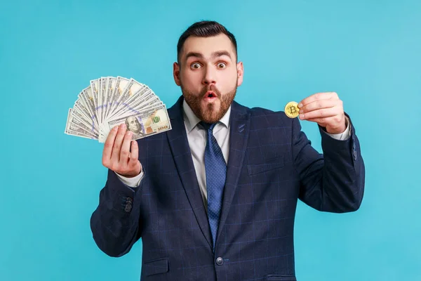 年轻的大胡子男子身穿官方风格的西装 脸上带着惊讶的表情 在镜头前展示美元钞票和黄金比特币的肖像 室内拍摄被蓝色背景隔离 — 图库照片