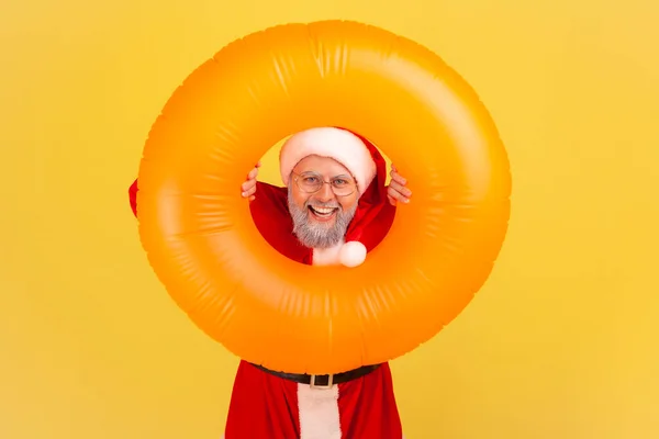 Smiling elderly man with gray beard in santa claus costume holding orange rubber ring in hands, looking at camera with happy expression, winter tour. Indoor studio shot isolated on yellow background.