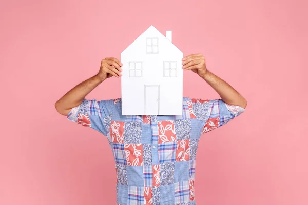 Unknown male person in blue casual style shirt covering his face with big paper house, dreams about own accommodation, estate agent. Indoor studio shot isolated on pink background.