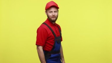 Kendine güveni tam bir işçi kameraya döner ve elleri kalçasında durur, kendine güveni tam, mavi üniformalı kırmızı tişört ve güneşlik şapkası vardır. Sarı arka planda izole edilmiş kapalı stüdyo çekimi.