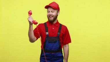 Geçmişe dönük sabit telefondan konuşan, mavi üniformalı, kırmızı tişörtlü ve vizör şapkalı yakışıklı çağrı merkezi çalışanı. Sarı arka planda izole edilmiş kapalı stüdyo çekimi.