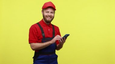Mutlu pozitif işçi akıllı telefon ekranını işaret ediyor ve mutlu bir şekilde gülümsüyor, mavi üniforma, tişört ve güneşlik takan bir uygulama tavsiye ediyor. Sarı arka planda izole edilmiş kapalı stüdyo çekimi.