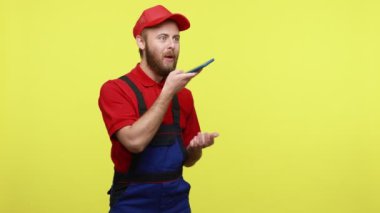 Akıllı telefon kullanan sakallı bir adam, müşteriye sesli mesaj ya da komut kaydeden, mavi üniformalı, kırmızı tişörtlü ve vizör şapkalı. Sarı arka planda izole edilmiş kapalı stüdyo çekimi.