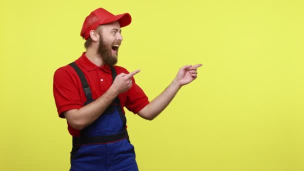 兴奋的男工把手指放在一边 展示广告或促销的复制空间 身穿蓝色制服 红色T恤和帽衫 在黄色背景下被隔离的室内拍摄 — 图库视频影像