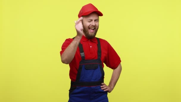 笑顔幸せな労働者の男性は 正の式でカメラに指を指して あなたを選択し 青い制服を着て 赤いTシャツとバイザーキャップ 黄色の背景に隔離された屋内スタジオショット — ストック動画