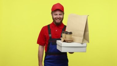 Elinde yiyecek ve kahve olan gülen kurye siparişler için kırmızı retro telefonu gösteriyor, mavi üniforma, tişört ve güneşlik takıyor. Sarı arka planda izole edilmiş kapalı stüdyo çekimi.