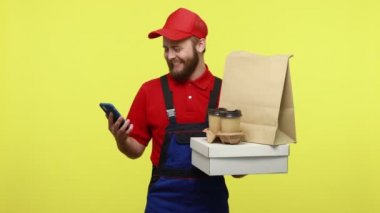Elinde yiyecek ve kahve paketiyle gülen kurye akıllı telefonlu müşteriyle konuşuyor mavi üniformalı, kırmızı tişörtlü ve güneşlik şapkalı. Sarı arka planda izole edilmiş kapalı stüdyo çekimi.