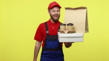 İyimser teslimatçı pakette yiyecek ve kahve tutuyor. Temassız ödemeler için terminali gösteriyor. Mavi üniforma, kırmızı tişört ve güneşlik takıyor. Sarı arka planda izole edilmiş kapalı stüdyo çekimi