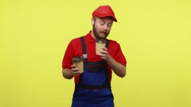 Uykulu zanaatkar esniyor, enerji toplamak için kahve içiyor, uykuya dalıyor, yorgun görünüyor, mavi üniforma, kırmızı tişört ve güneşlik takıyor. Sarı arka planda izole edilmiş kapalı stüdyo çekimi.