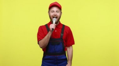 Şirket partisinde eğlenen, mavi üniformalı, tişörtlü ve güneşlik şapkalı, iş arkadaşına şarkı söylemesi için mikrofon sunan becerikli bir adam. Sarı arka planda izole edilmiş kapalı stüdyo çekimi.
