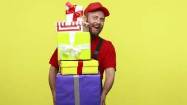 İyimser teslimatçı elinde hediyeler, paketlenmiş hediye kutularının arkasına saklanıyor, mavi üniforma, kırmızı tişört ve güneşlik takıyor. Sarı arka planda izole edilmiş kapalı stüdyo çekimi.