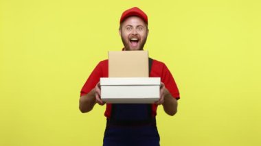 Müşteriye karton paketler teslim etmek için heyecanlı kurye, hızlı profesyonel teslimat servisi, mavi üniforma, kırmızı tişört ve güneşlik takıyor. Sarı arka planda izole edilmiş kapalı stüdyo çekimi.