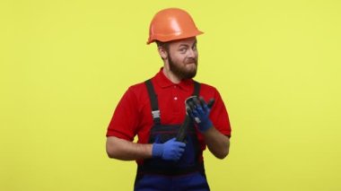 İyimser müteahhit ayarlanabilir İngiliz anahtarı tutuyor ve kameraya gülümsüyor mavi tulum, kırmızı tişört ve turuncu başlık giyiyor. Sarı arka planda izole edilmiş kapalı stüdyo çekimi.