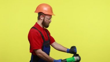 Marangoz ya da inşaatçının yan görünüş portresi testereyle çalışıyor, inşaat için testereyi kesiyor, mavi tulum ve turuncu kask giyiyor. Sarı arka planda izole edilmiş kapalı stüdyo çekimi.