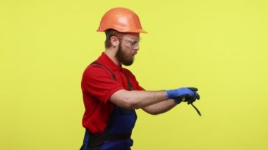 Ciddi bir müteahhitin bir şeyleri ölçtüğü, konsantre bir ifadeye sahip olduğu, mavi tulum giydiği ve turuncu kask taktığı yan görüş. Sarı arka planda izole edilmiş kapalı stüdyo çekimi.