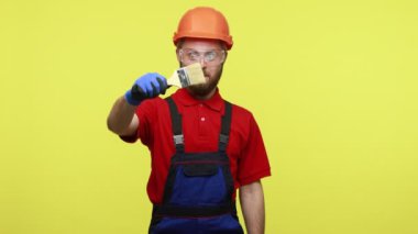 İyimser mimar ya da ressam elinde boya fırçası tutarken çalışıyormuş gibi yapıyor, kameraya bakıyor, mavi tulum ve turuncu kask takıyor. Sarı arka planda izole edilmiş kapalı stüdyo çekimi.
