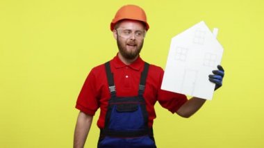 Elinde kağıt anahtar ve ev tutan gülümseyen müteahhit, inşaatın bittiğini duyurdu, mavi tulum, kırmızı tişört ve turuncu başlık giydi. Sarı arka planda izole edilmiş kapalı stüdyo çekimi.