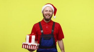 Elinde hediyeler, alışveriş poşetleri, hızlı teslimat, mavi tulum ve Noel Baba şapkası olan mutlu teslimatçı adam. Sarı arka planda izole edilmiş kapalı stüdyo çekimi.