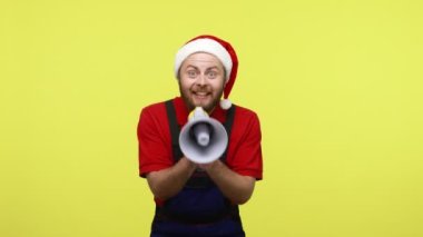 Heyecanlı işçi megafonla bağırıyor, Noel indirimleri ilan ediyor, mavi tulum ve Noel Baba şapkası takmış heyecanlı görünüyor. Sarı arka planda izole edilmiş kapalı stüdyo çekimi.