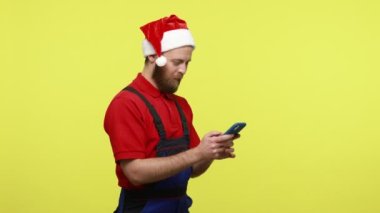 Akıllı telefon kullanan, yüzünde olumlu ifade olan mavi tulum ve Noel Baba şapkası takan işçilerin yan görünüm portresi. Sarı arka planda izole edilmiş kapalı stüdyo çekimi.