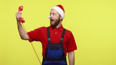 Çağrı merkezinin heyecanlı operatörü kırmızı sabit telefon ve kameraya işaret veriyor, iyimserliğini ifade ediyor, mavi tulum giyiyor ve Noel Baba şapkası takıyor. Sarı arka planda izole edilmiş kapalı stüdyo çekimi.