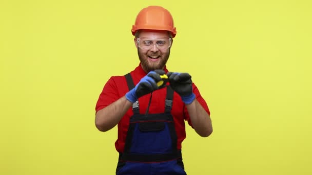 快乐的积极的建设者 双手拿着黄色的尺码带子站在那里 面部表情滑稽 身穿蓝色工作服 头戴橙色头盔 在黄色背景下被隔离的室内拍摄 — 图库视频影像