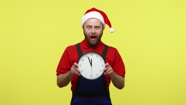 時計を見せ 青い全体 赤いTシャツとサンタのクラスの帽子を身に着けて 大晦日まで仕事を終えるのが嬉しい幸せな正の男 黄色の背景に隔離された屋内スタジオショット — ストック動画