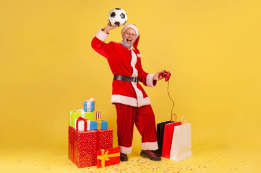 Gri sakallı, Noel Baba kostümlü, elinde futbol topuyla kol kaldıran, elinde joystickle futbol video oyununda kazandığı zaferi kutlayan bir adam. Sarı arka planda kapalı stüdyo çekimleri.