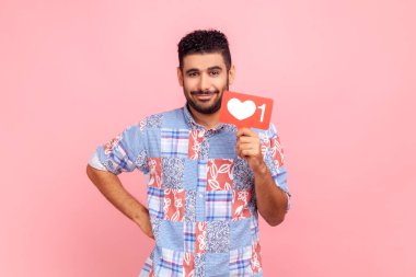 İlginç içeriği takip et! Mavi tişört giyen, elinde sosyal medya kalp ikonu tutan, gülümseyen sakallı bir adamın portresi, elini havada tutuyor. Pembe arka planda kapalı stüdyo çekimleri.