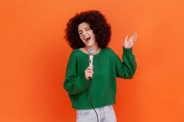 手にマイクで立って緑のカジュアルなスタイルのセーターを着てアフロの髪型と興奮した女性の肖像 幸せな表情で曲を歌う オレンジの背景に隔離された屋内スタジオショット — ストック写真