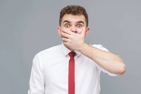 Portrait of young shocked man in white shirt and tie standing closing his mouth with hand and looking at camera with shocked face and big eyes. indoor isolated on gray background.