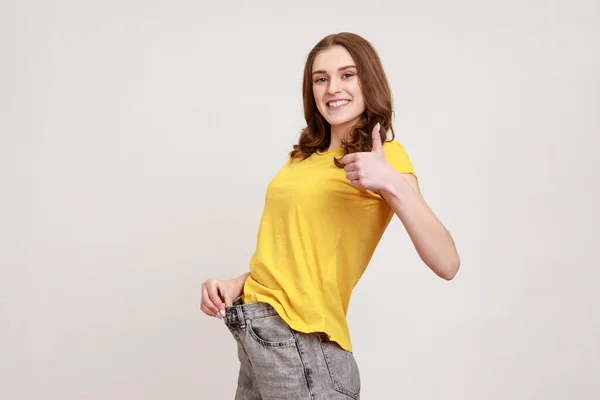 美しい幸せなティーンエイジャーの女の子のプロフィール肖像画は 大きなズボンでスリムな腰を示し 親指を表示する意志力を持っています 成功した減量 灰色の背景に隔離された屋内スタジオショット — ストック写真
