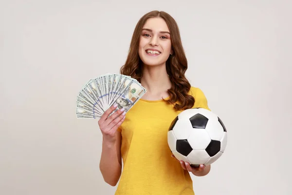 黄色のTシャツ保持サッカーボールとドルのファンでおもちゃの笑顔で十代の女の子 サッカー選手権での勝利を祝います 灰色の背景に隔離された屋内スタジオショット — ストック写真